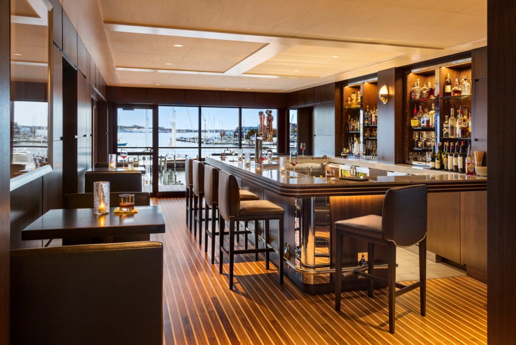 Warmly lit bar interior with golden-brown striped wood flooring, dark seating and waterfront windows