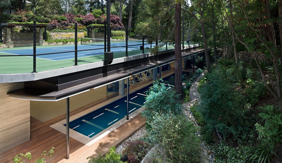 A long deck and pool sit beside dense planting and beneath a glazed sports court