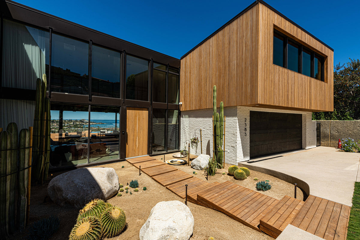A wood walkway rises across a planted front garden toward a modern timber-clad house
