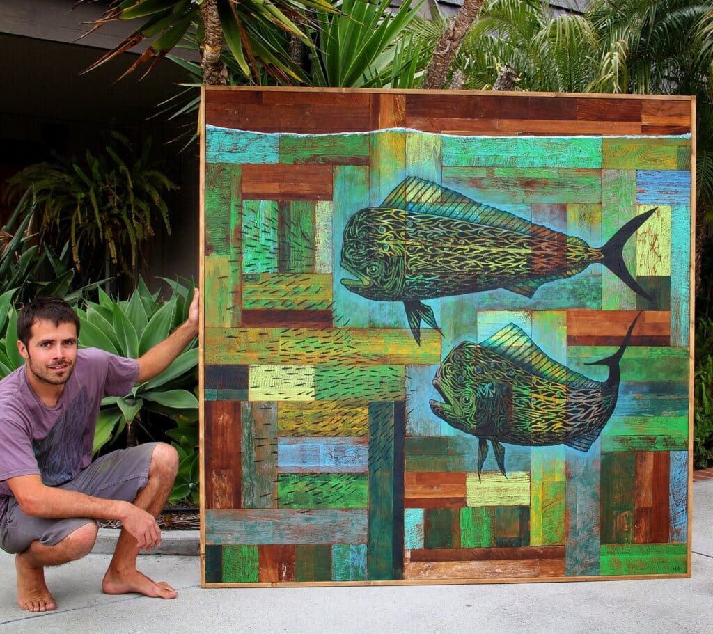 Artist beside a large colorful fish artwork made on a wood panel